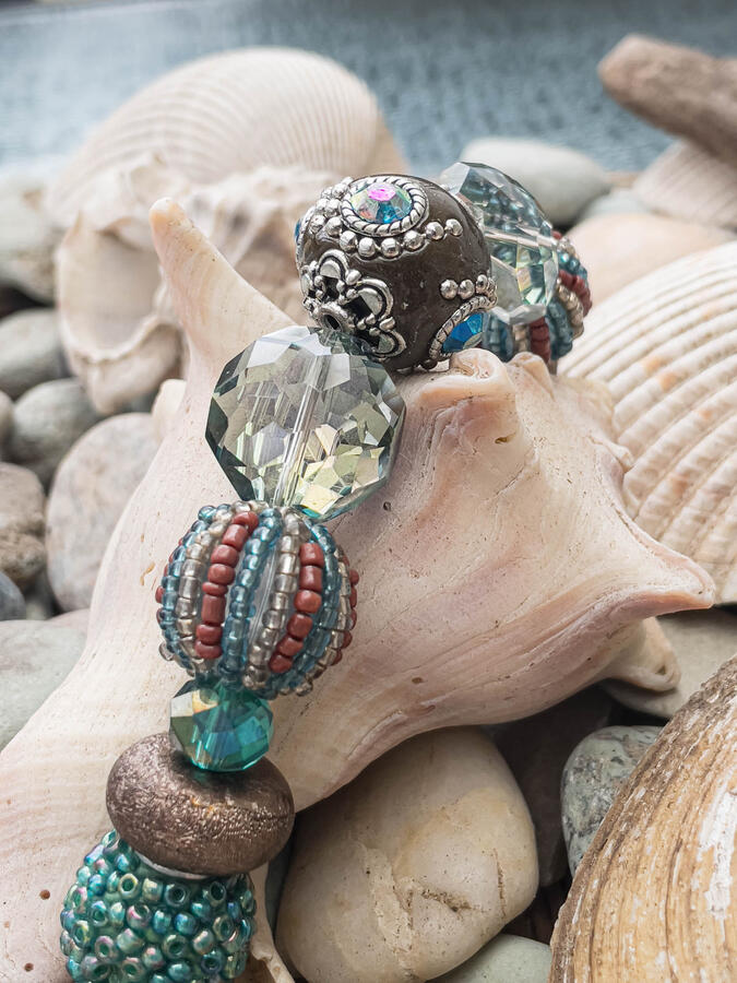 Handcrafted artisan bracelet featuring an ornate silver-capped dark bead, faceted smokey crystals, and textured seed bead spheres, draped over a large natural conch shell and sea stones.