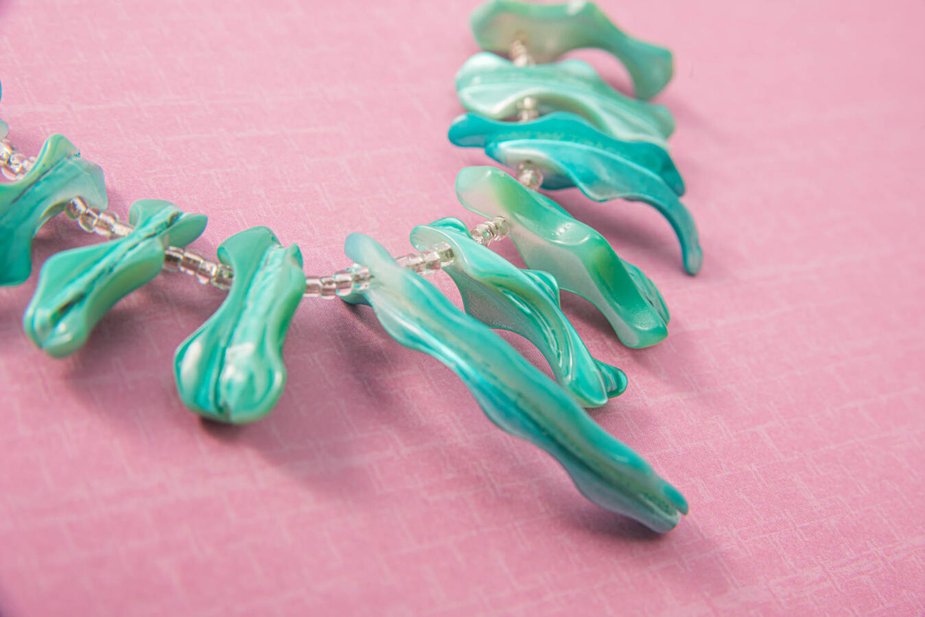 A handcrafted necklace featuring long, organic-shaped iridescent turquoise mother-of-pearl spires on a textured bright pink background.