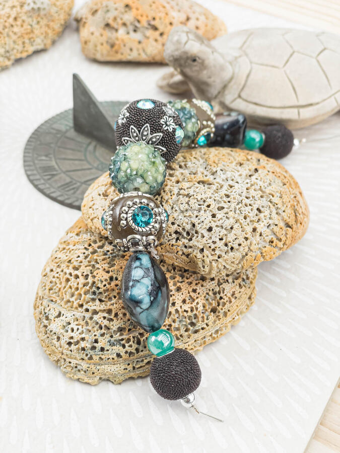 Handcrafted artisan bracelet with ornate silver-capped beads, turquoise stones, and brown textured accents displayed on sea coral next to a vintage sundial and stone turtle.