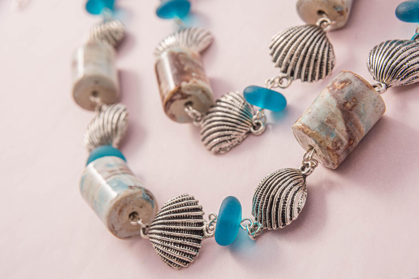 Close-up of a handcrafted silver necklace featuring detailed scallop shell charms, cylindrical marbled earth-tone beads, and translucent teal sea glass accents.