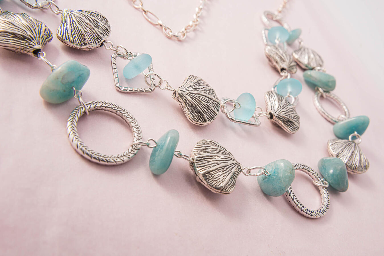 A handcrafted multi-layered silver necklace featuring detailed scallop shell charms, smooth turquoise-colored stones, and translucent light blue sea glass beads on a soft pink background.
