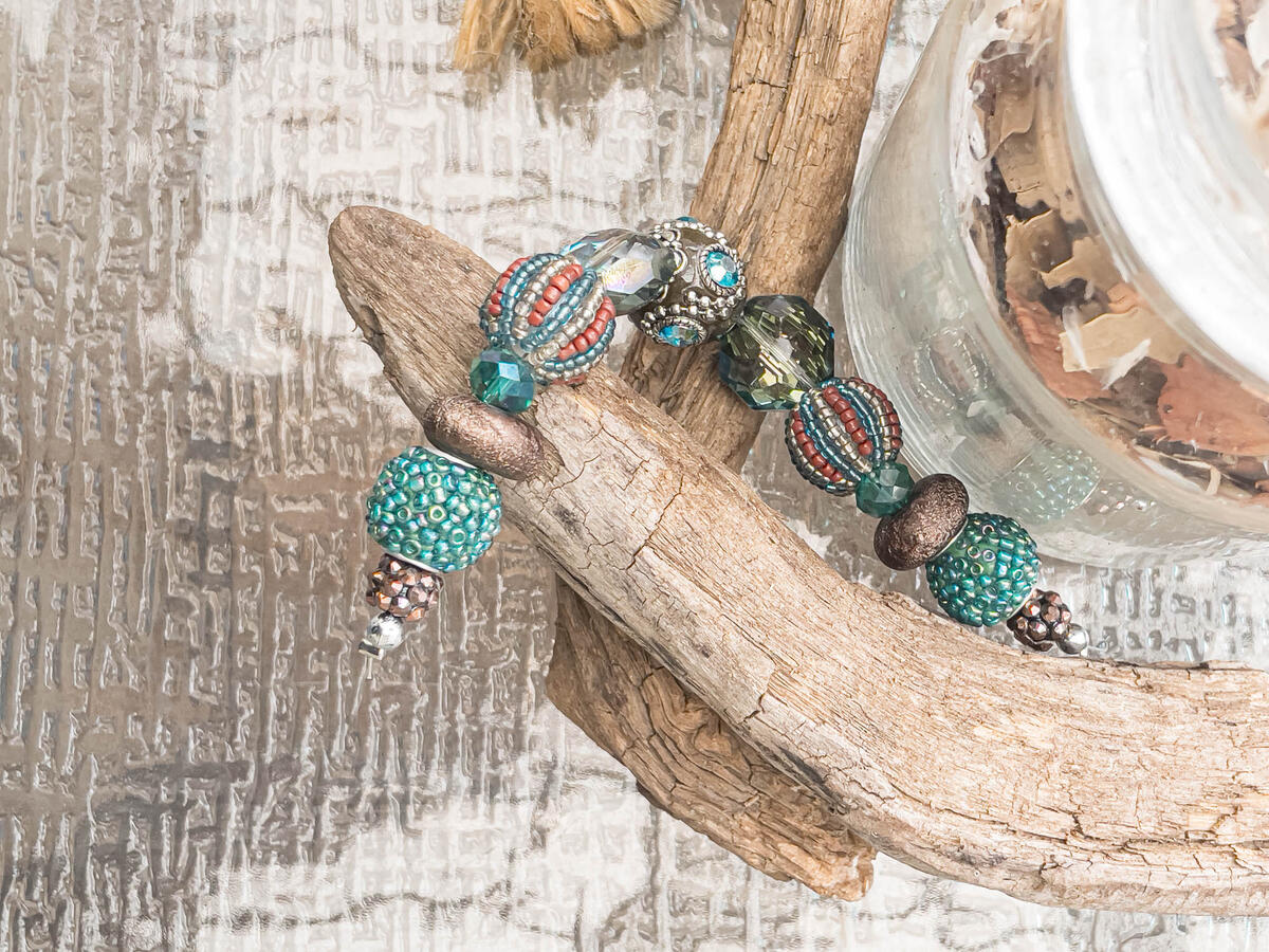 A handcrafted bohemian-style beaded bracelet featuring teal, bronze, and iridescent crystal beads draped over natural driftwood.