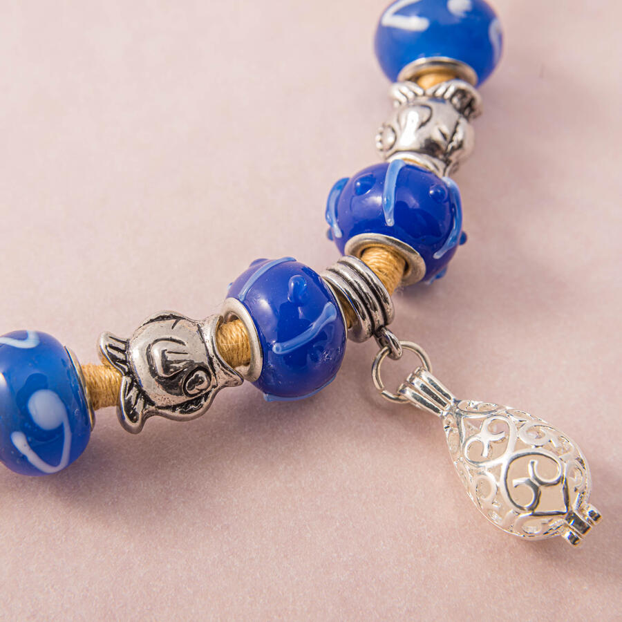 Close-up of a handcrafted necklace featuring bold blue glass beads with white swirl patterns, silver fish-shaped spacers, and a silver filigree cage pendant on a tan cord.