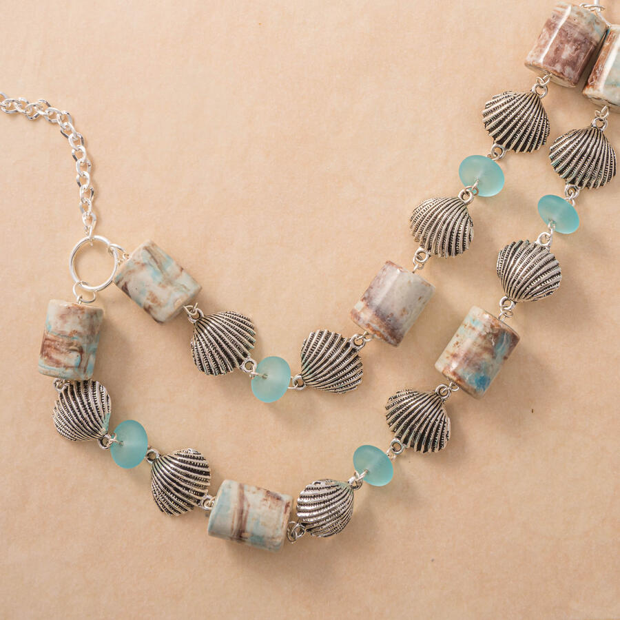 A detailed view of a silver necklace featuring ornate scallop shell charms, marbled tan and blue cylindrical stones, and translucent teal glass beads on a neutral background.
