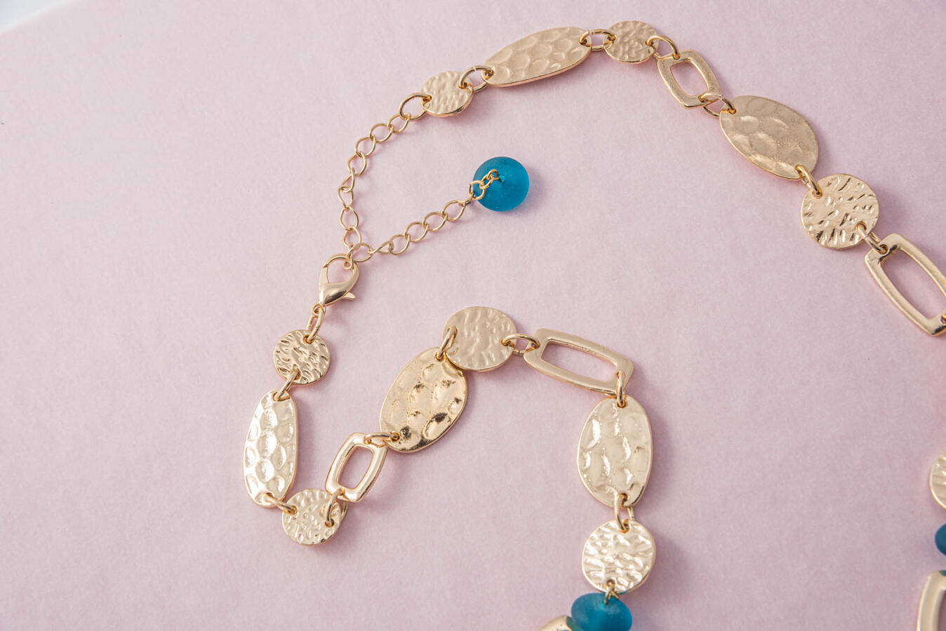 A handcrafted gold-toned necklace featuring hammered coin-style links and vibrant teal glass beads on a soft pink background.