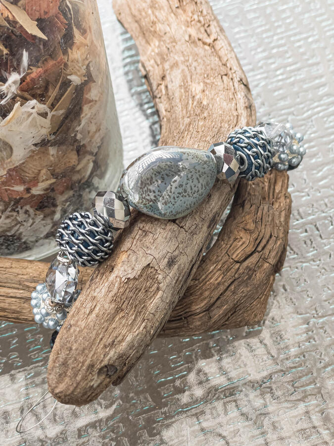 Handcrafted artisan bracelet featuring a large, polished grey speckled stone, silver chain-wrapped blue beads, and faceted silver crystals displayed on a piece of weathered driftwood.