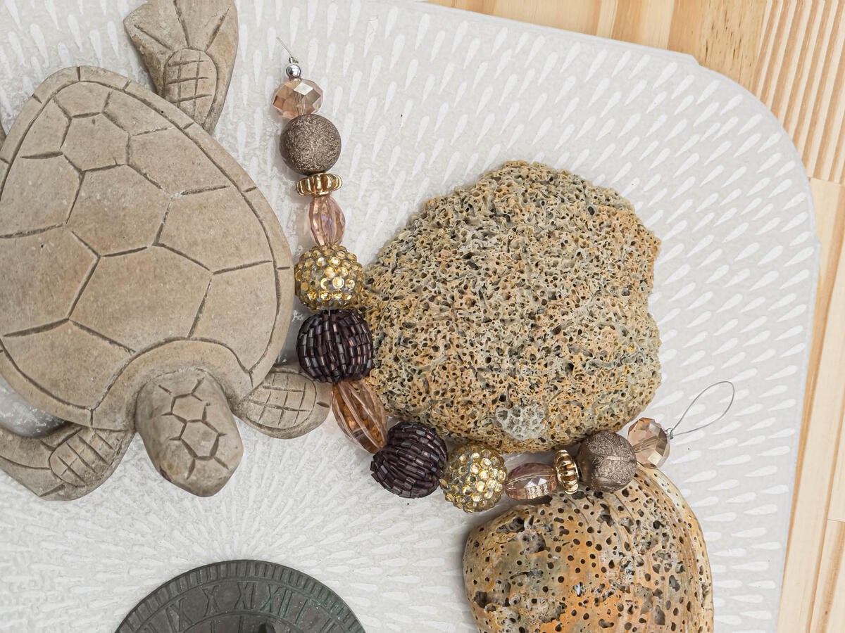 Handcrafted artisan bracelet featuring textured bronze beads, gold-encrusted crystal spheres, and translucent champagne accents, displayed on a textured stone surface with a stone turtle and vintage sundial.