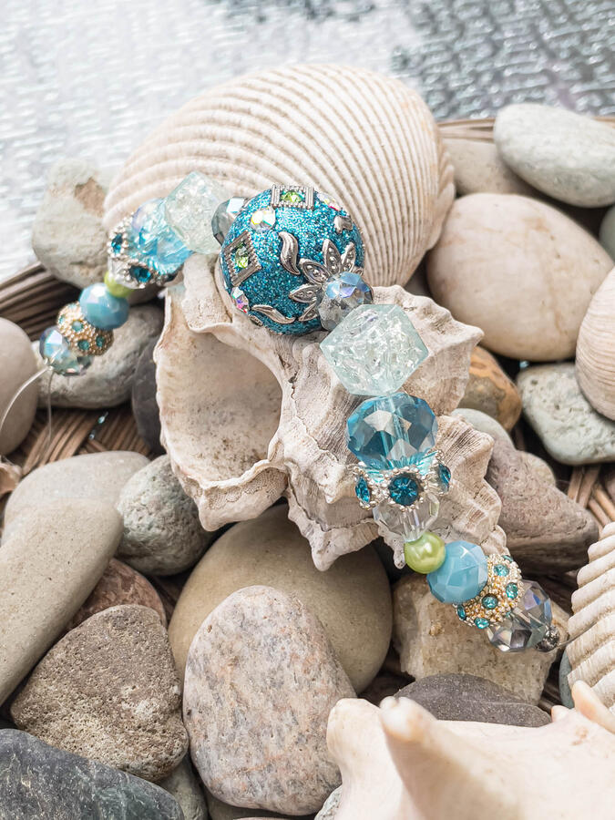 Intricate aqua blue and silver beaded bracelet with floral crystal accents, draped over a murex seashell and smooth river rocks.
